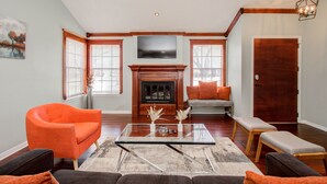 A living room with a fireplace, a television mounted above it, and a large window with blinds. - The Greenhouse (Houston)