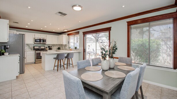 A modern kitchen with white cabinets, a stainless steel refrigerator, and a built-in oven. There is a dining area with a wooden table and grey chairs. - The Greenhouse (Houston)
