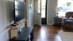 Ein Wohnzimmer mit einem Flachbildfernseher auf einem Holzständer, einem kleinen Tisch mit einer Lampe, einem Sessel und einem großen Fenster mit Blick ins Freie.