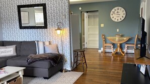 Ein Wohnzimmer mit grauem Sofa, einem kleinen Couchtisch, einem gemusterten Teppich, Holzboden, einem Essbereich mit Holztisch und Stühlen, einer Wanduhr und einem Deckenventilator.