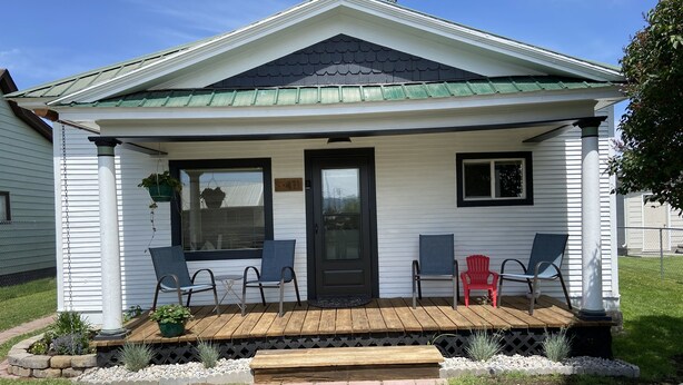 Ein kleines Haus mit Veranda, zwei Stühle, ein roter Sessel und ein Blumentopf.