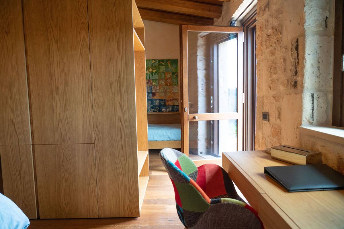 A room with a wooden wardrobe, a bed with a blue mattress, a colorful chair, a wooden desk with a laptop, and a window with a view of a garden.