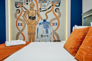 A hotel room with a bed featuring orange bedding, a wall mural of Star Wars characters, and a window with a view of the sky.