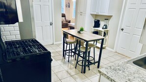 Une cuisine équipée d’une cuisinière à gaz, d’une poubelle noire, d’une table en bois avec deux chaises et d’un plan de travail avec un évier.