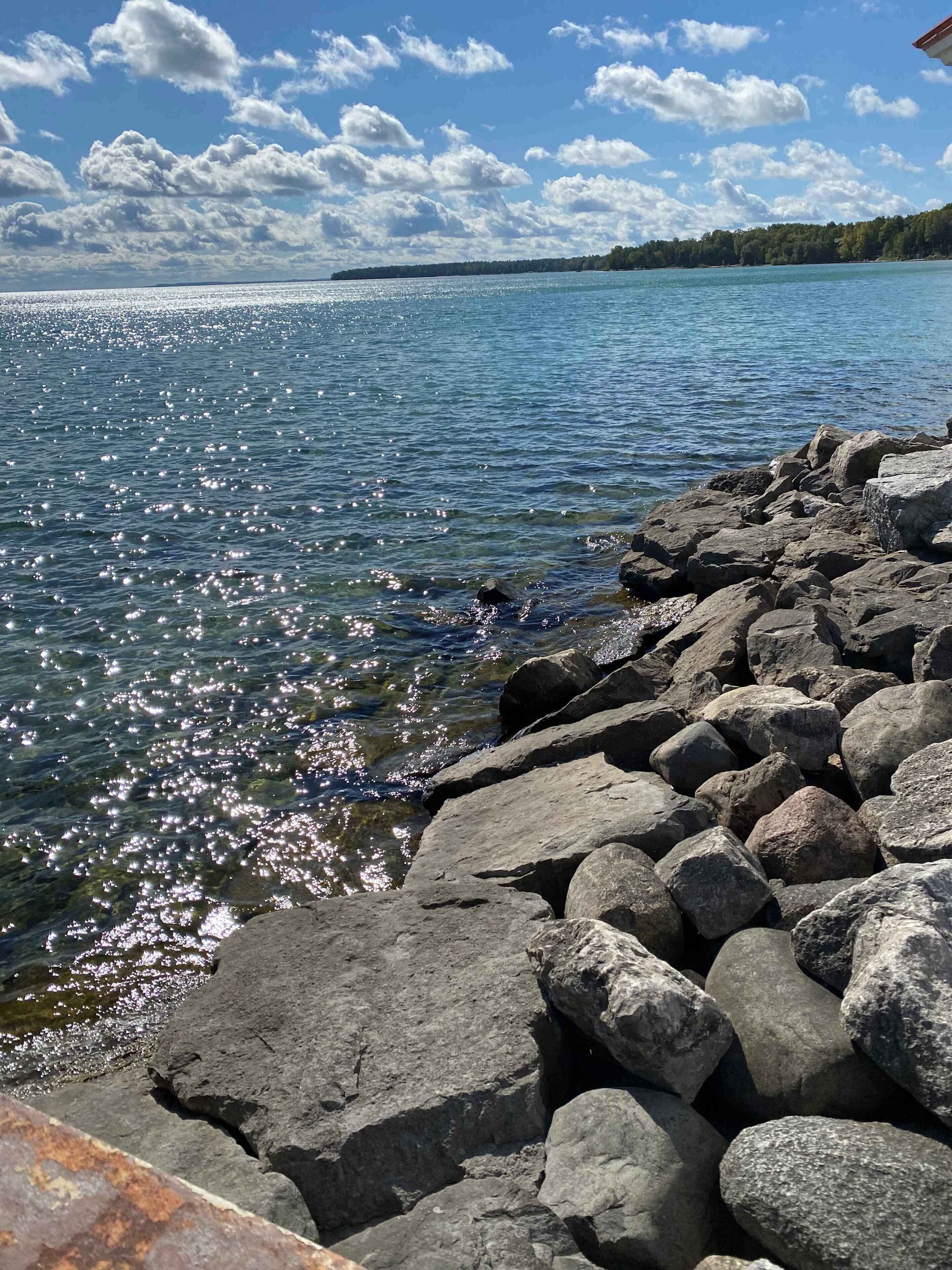 Un rivage rocheux bordé d’une eau claire qui reflète la lumière du soleil, une ligne d’arbres à l’horizon et un ciel partiellement nuageux.