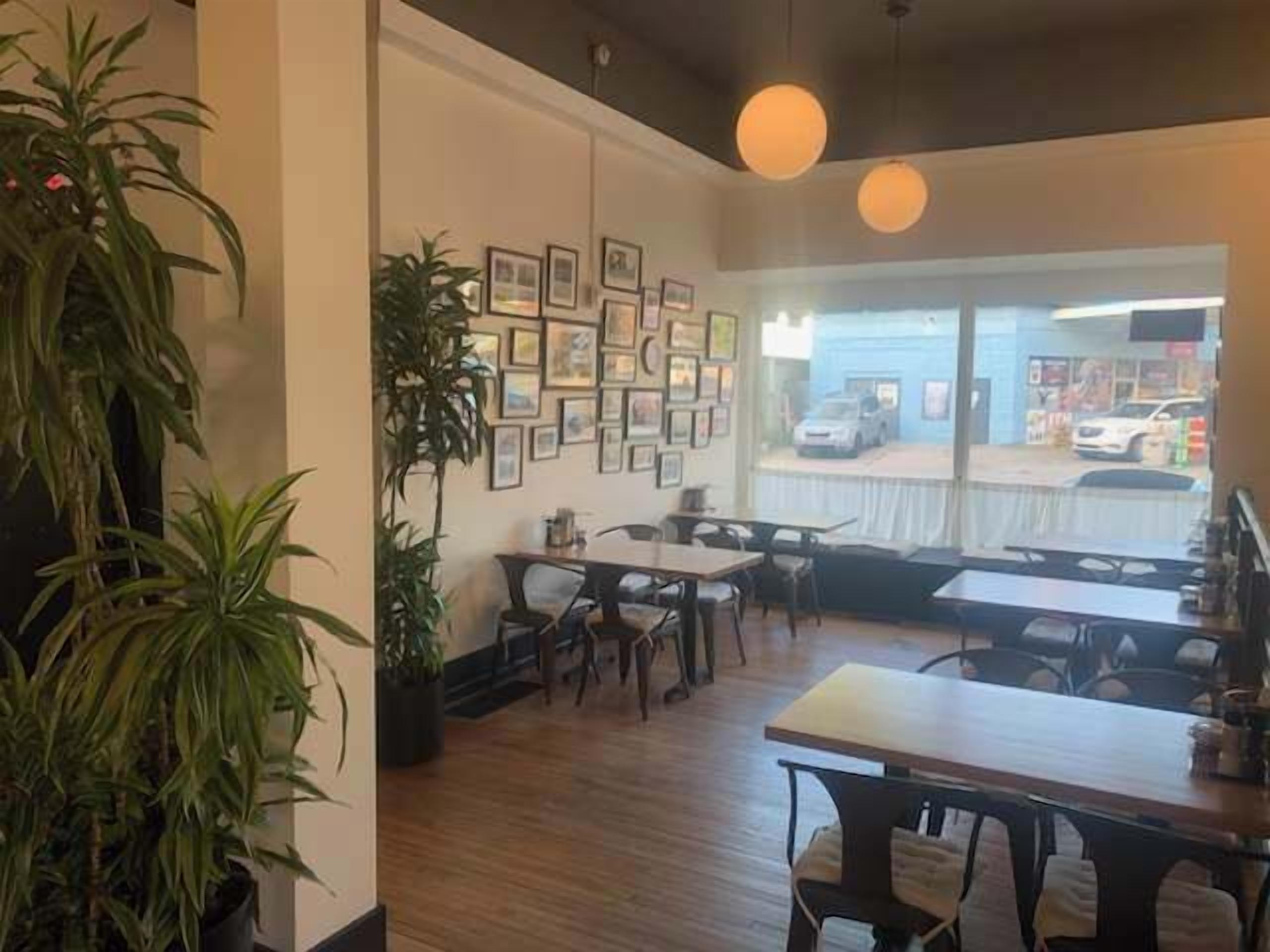 A restaurant interior with wooden tables, chairs, potted plants, framed pictures on the wall, and a view of the street outside.