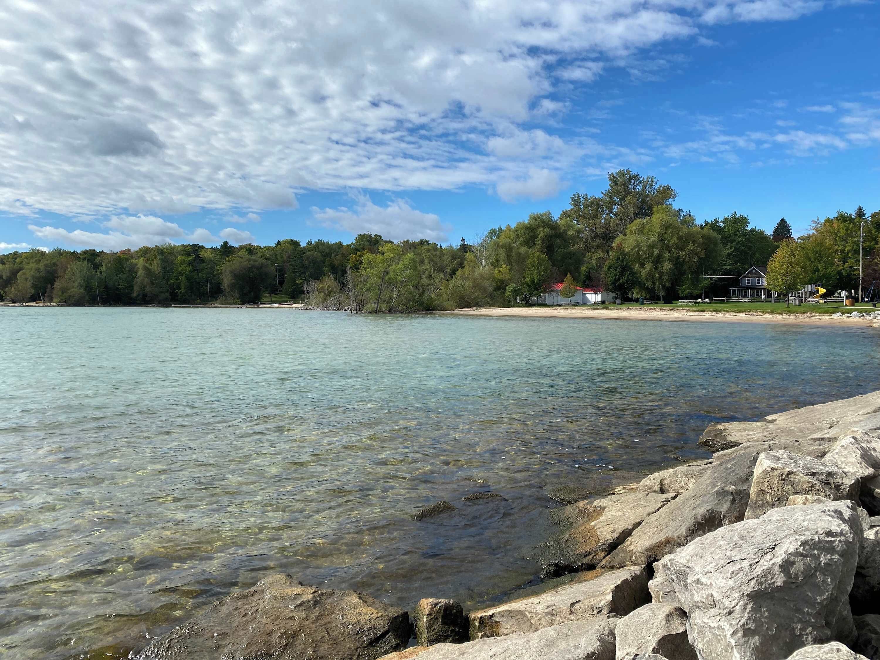 A clear lake with visible rocks in the foreground, a sandy beach, and a forested area with buildings in the background.