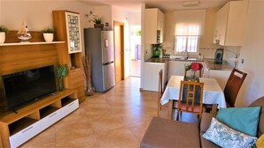 Un espace de vie compact comprenant un téléviseur, un meuble de rangement en bois, un coin repas avec une table et des chaises, une cuisine avec un réfrigérateur et une petite table à manger agrémentée d’un vase.