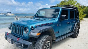 Un Jeep Rubicon blau estacionat a una platja sorrenca, amb un cos d'aigua i embarcacions al fons.