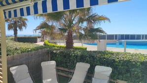 A covered patio with a view of a pool and beach, three white lounge chairs, and a striped awning. - Beachfront apartment with pool, air conditioning and wifi (Frontignan)