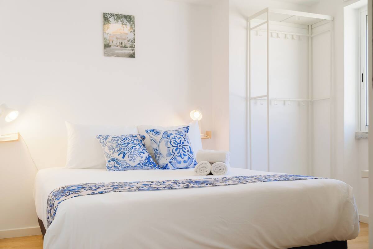 A neatly made bed with a white and blue patterned bedspread and pillows, a bedside lamp, and a wall-mounted picture.