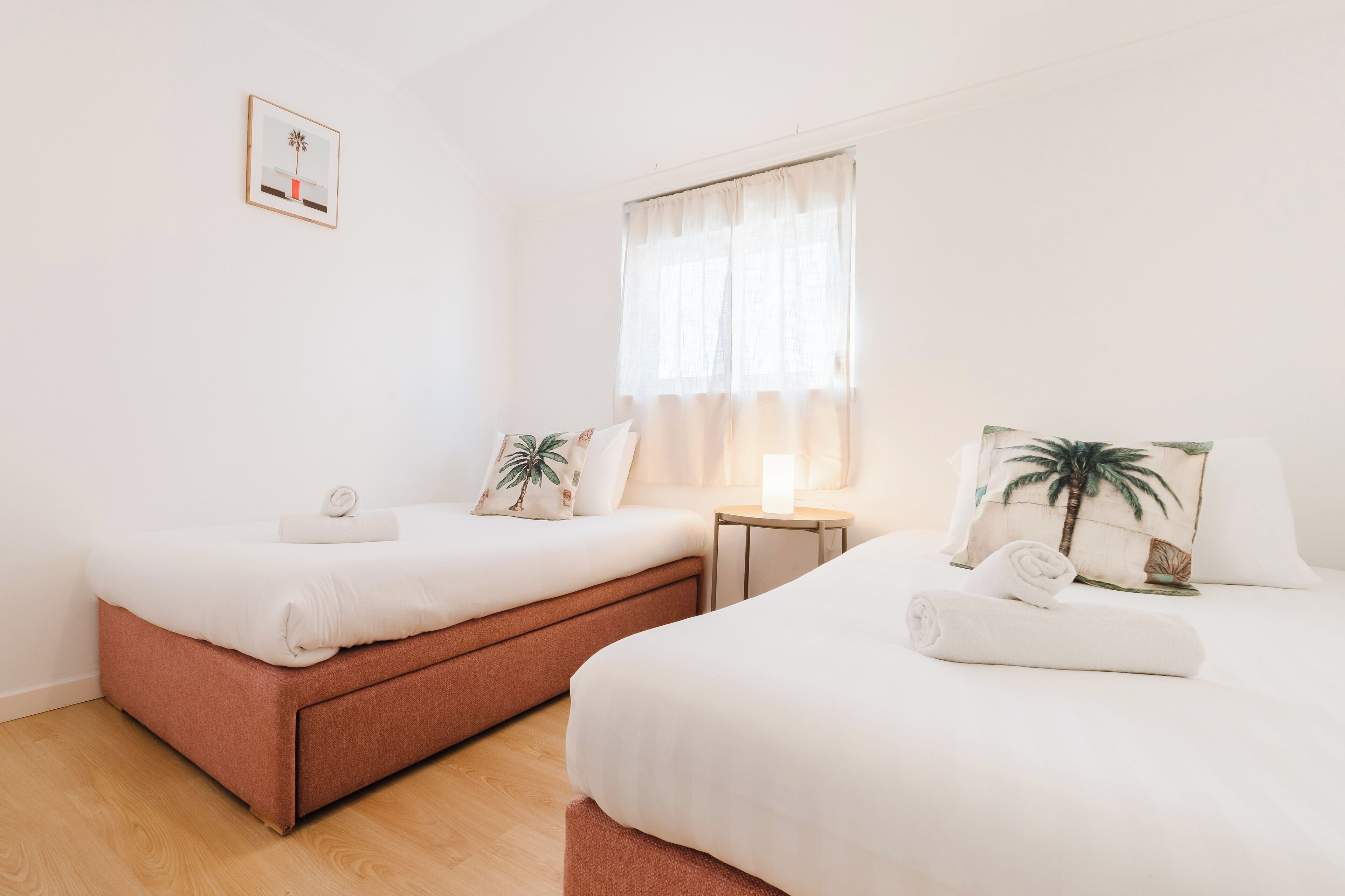 Habitación de hotel con dos camas, una mesita, una mesita de noche y una ventana con cortinas traslúcidas.