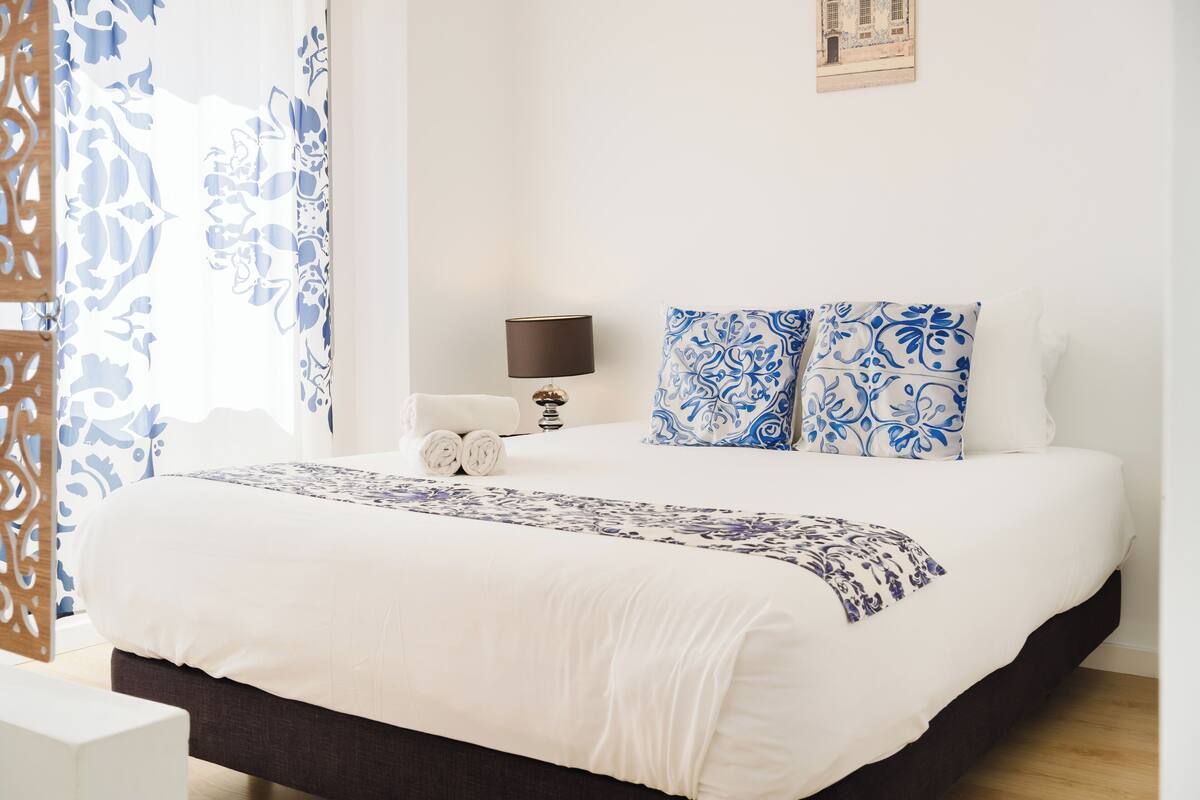 A neatly made bed with white and blue patterned bedding, a bedside table with a lamp, and a wall-mounted picture.