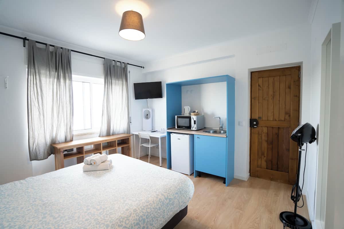 A compact hotel room with a bed, a desk, a small kitchenette, a television, and a wooden door.