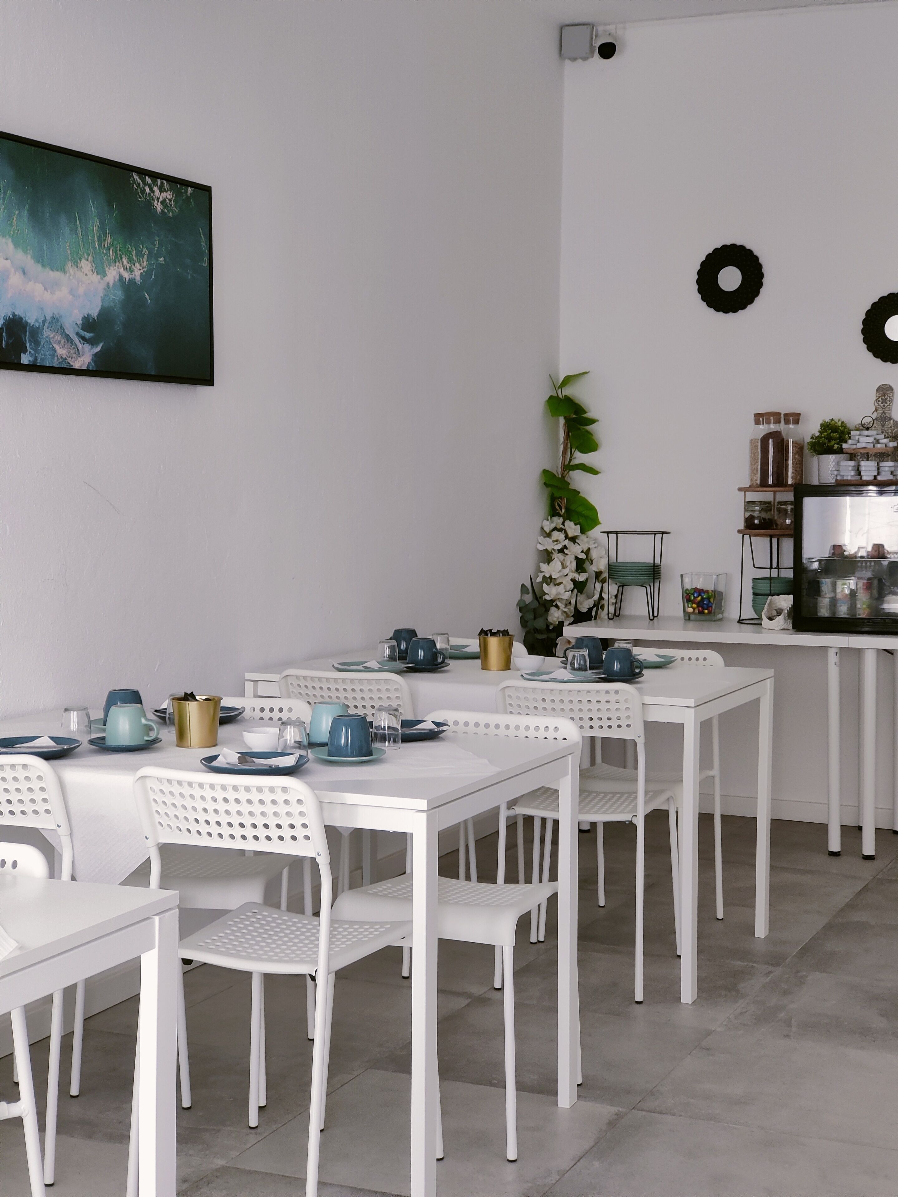 A dining area with white tables and chairs, a painting on the wall, and a shelf with decorative items.
