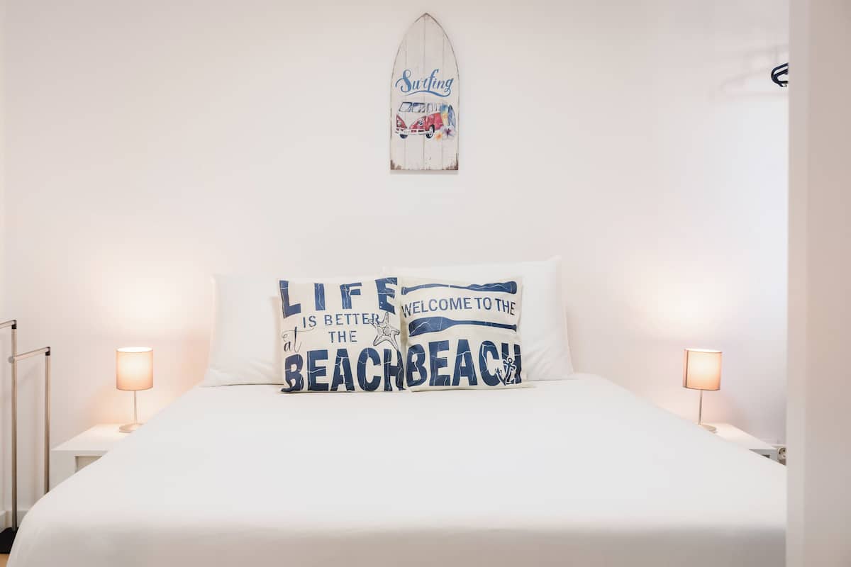 A bed with white bedding, two bedside lamps, and two decorative pillows with beach-themed text.