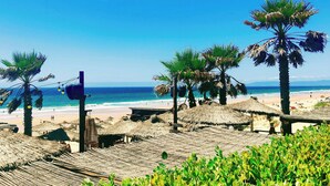 A beach with palm trees, thatched huts, and a clear blue sky. - Villa Paraiso Da Caparica (Almada)