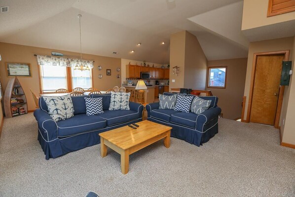 A living room with two blue sofas, a wooden coffee table, and a kitchen in the background. - Play All Day - 6BR Oceanside home - Short walk to beach, Private Pool, Dogs Allowed (Corolla)