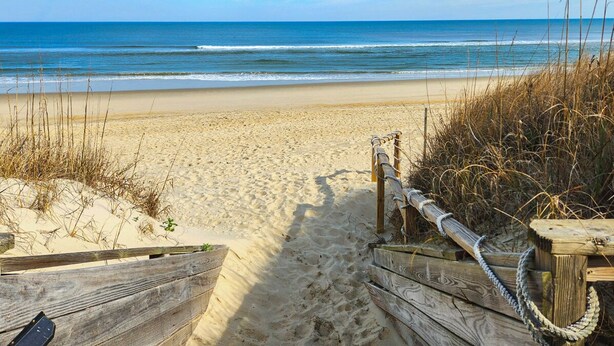Un sendero de madera bordeando la playa que lleva al océano, rodeado de arena y vegetación escasa.