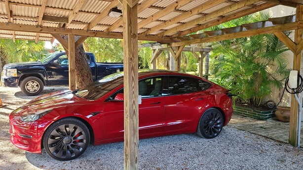 Une Tesla rouge est garée sous une pergola en bois, sur un sol recouvert de gravier, entourée de verdure et avec un camion noir en arrière-plan.