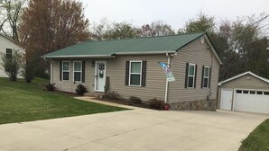 A single-story house with a green metal roof, a front door with a wreath, and a garage. - Adorable “Hudson Hideaway Cottage” (Hudson)