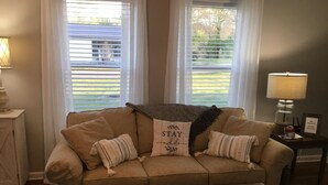 A living room with a beige sofa, two pillows, a throw blanket, a coffee table with a book and a pan, and a lamp on a side table. - Adorable “Hudson Hideaway Cottage” (Hudson)