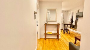 A narrow hallway with wooden flooring leading to a dining area with a table and chairs. A small shelf with books is visible, and a mirror hangs on the wall. - North End, Lil Italy, TD Garden (Boston)