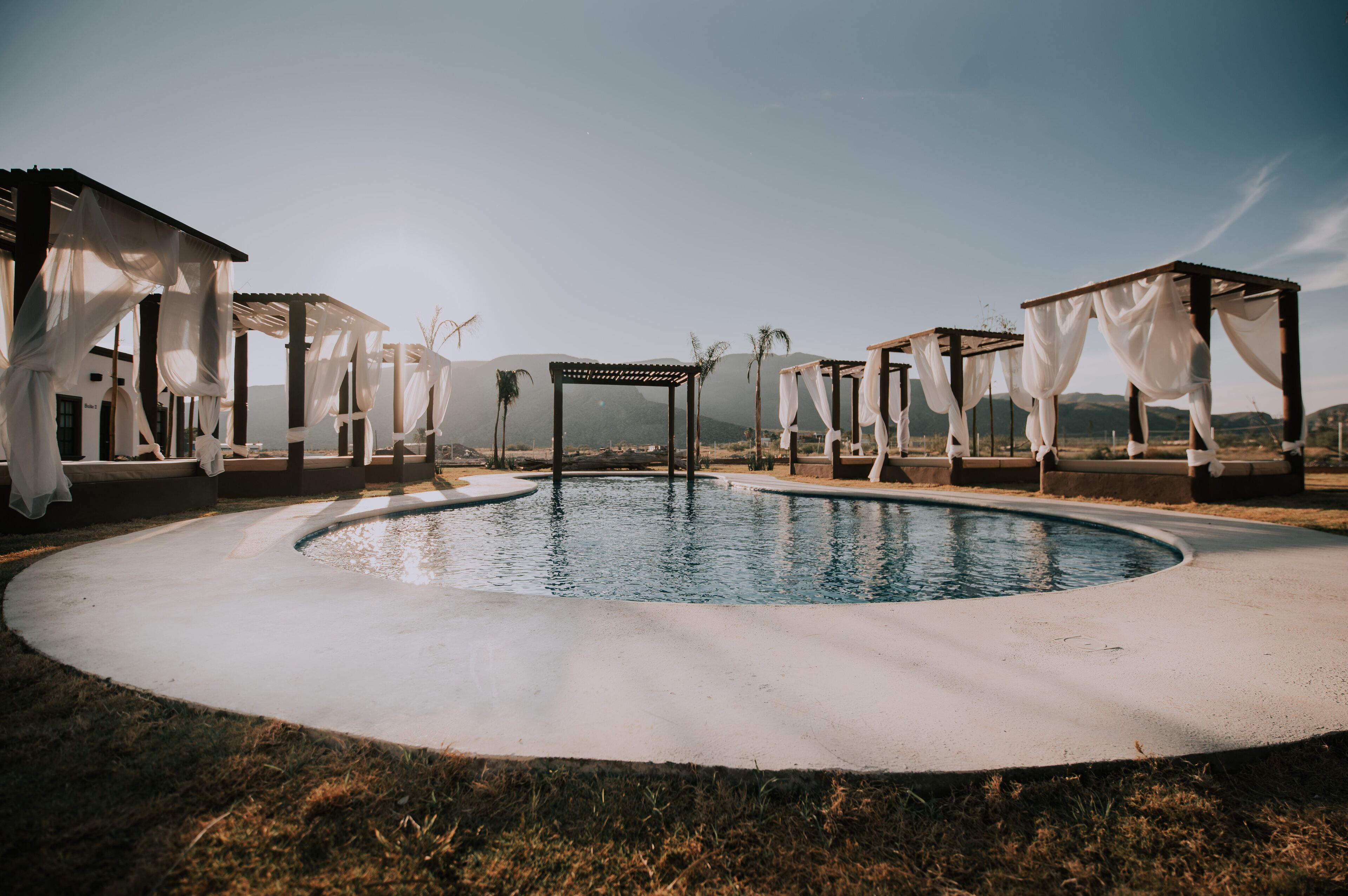 Zona de piscina con una estructura de pérgola, rodeada de toldos blancos y una vista despejada de montañas distantes.