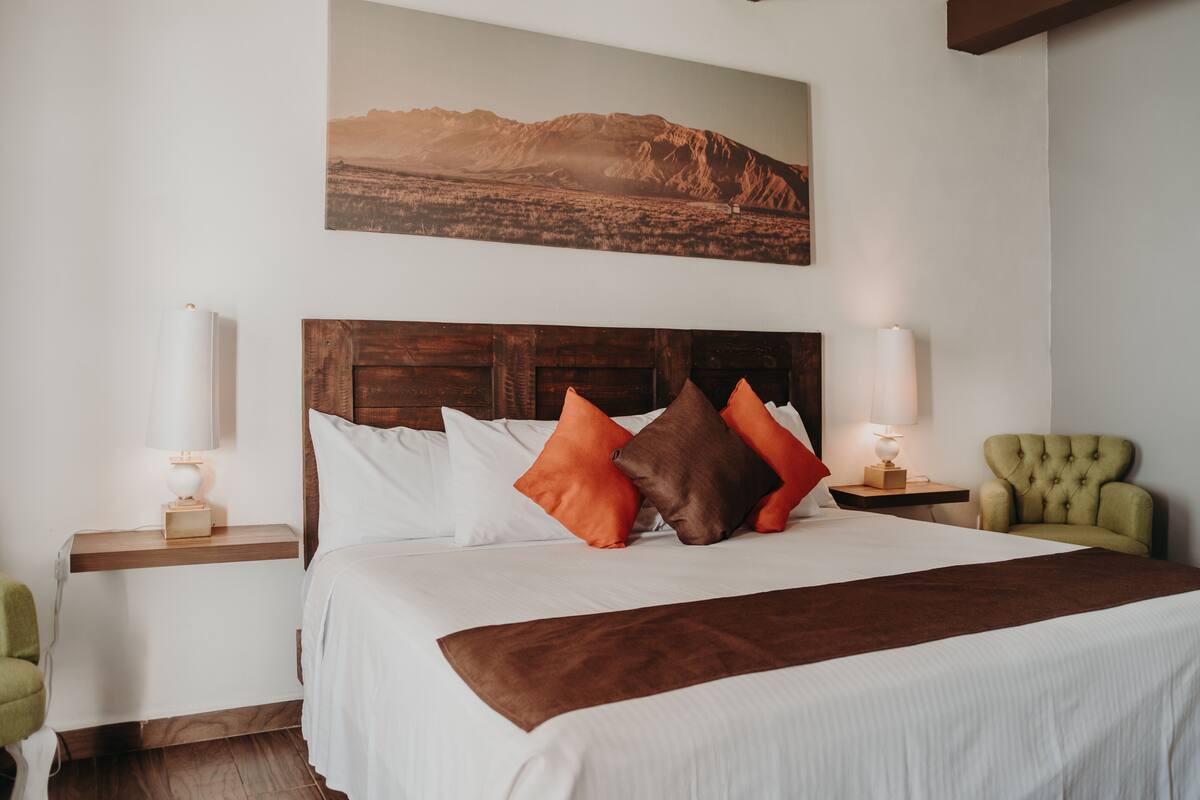 A neatly made bed with white and brown bedding, two bedside lamps, a green armchair, and a mountain landscape painting on the wall.