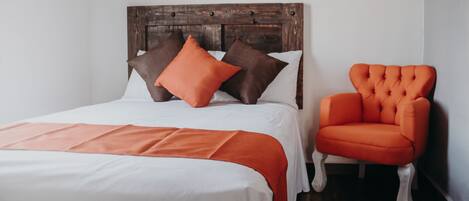 Una cama bien hecha con cabecera de madera, una silla naranja brillante y una colcha blanca.