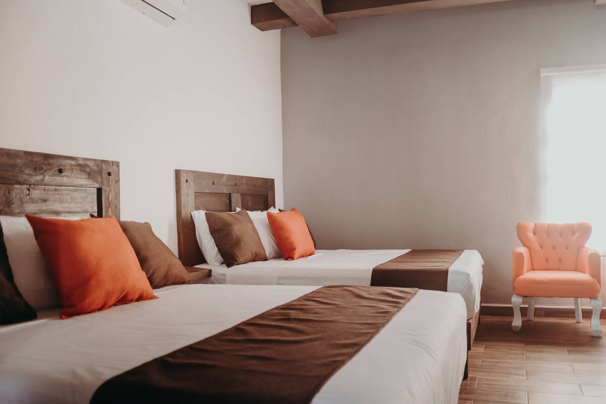 A hotel room with two beds, wooden headboards, orange and brown pillows, a wooden chair, and a large window.