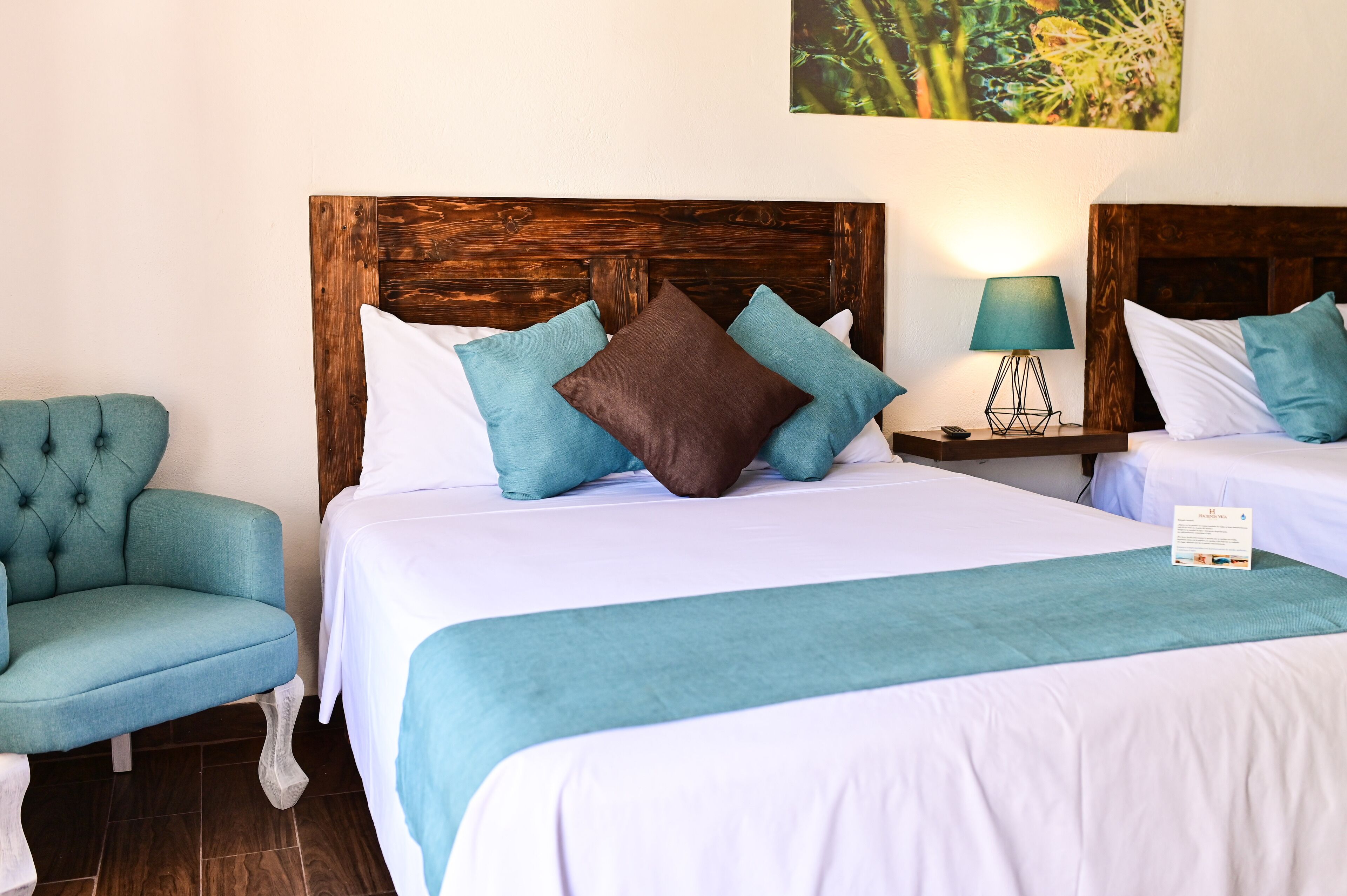 Quarto de hotel com duas camas, cabeceira de madeira, roupa de cama em tons de verde-água e branco, uma poltrona verde-água e uma mesa de cabeceira com abajur.