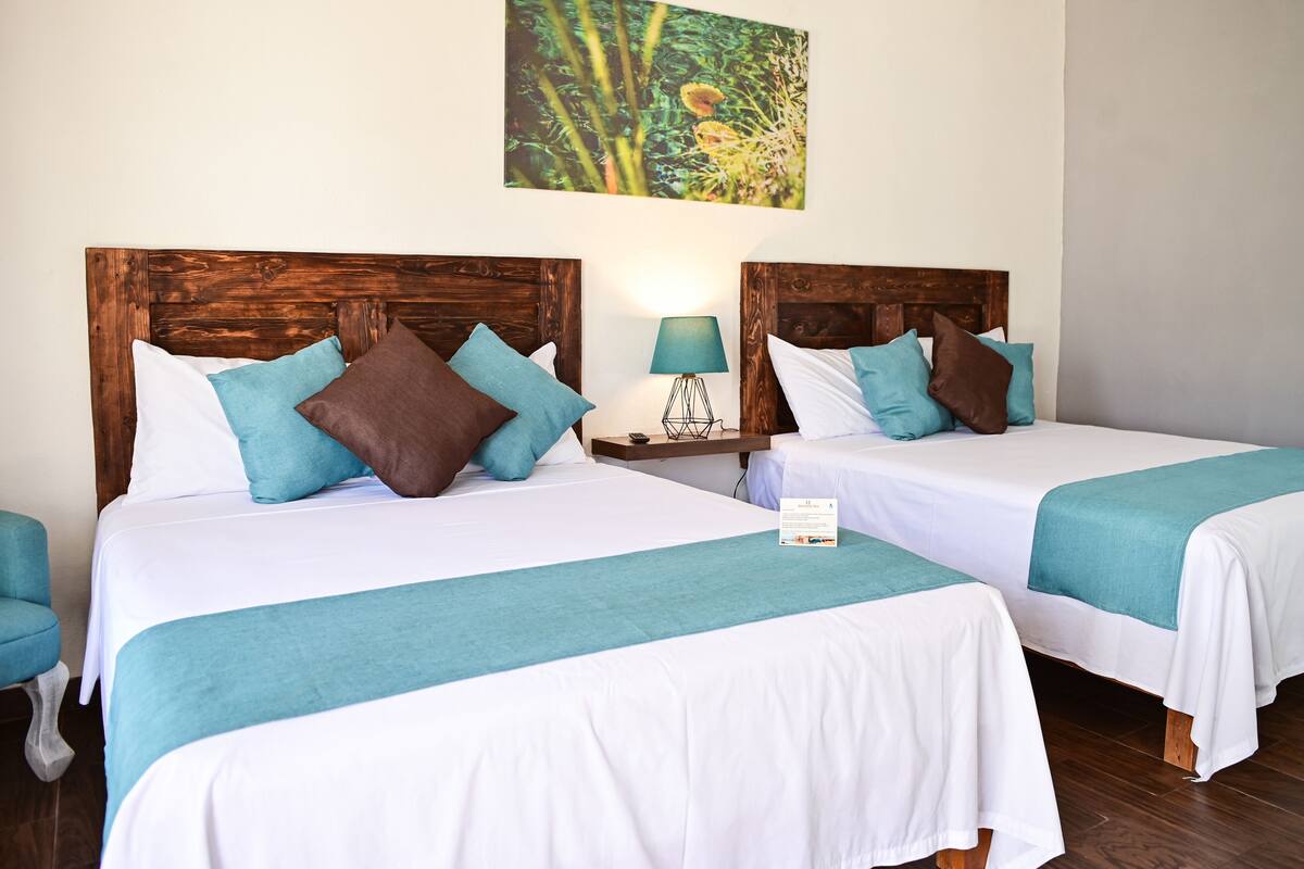 Two twin beds with white and teal bedding, a wooden headboard, a nightstand with a lamp, and a wall-mounted artwork.