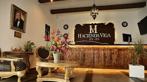 A reception area with a wooden reception desk, a framed portrait, a clock, and a potted plant. - Hacienda Viga 2020 Hotel (Cuatro Ciénegas)
