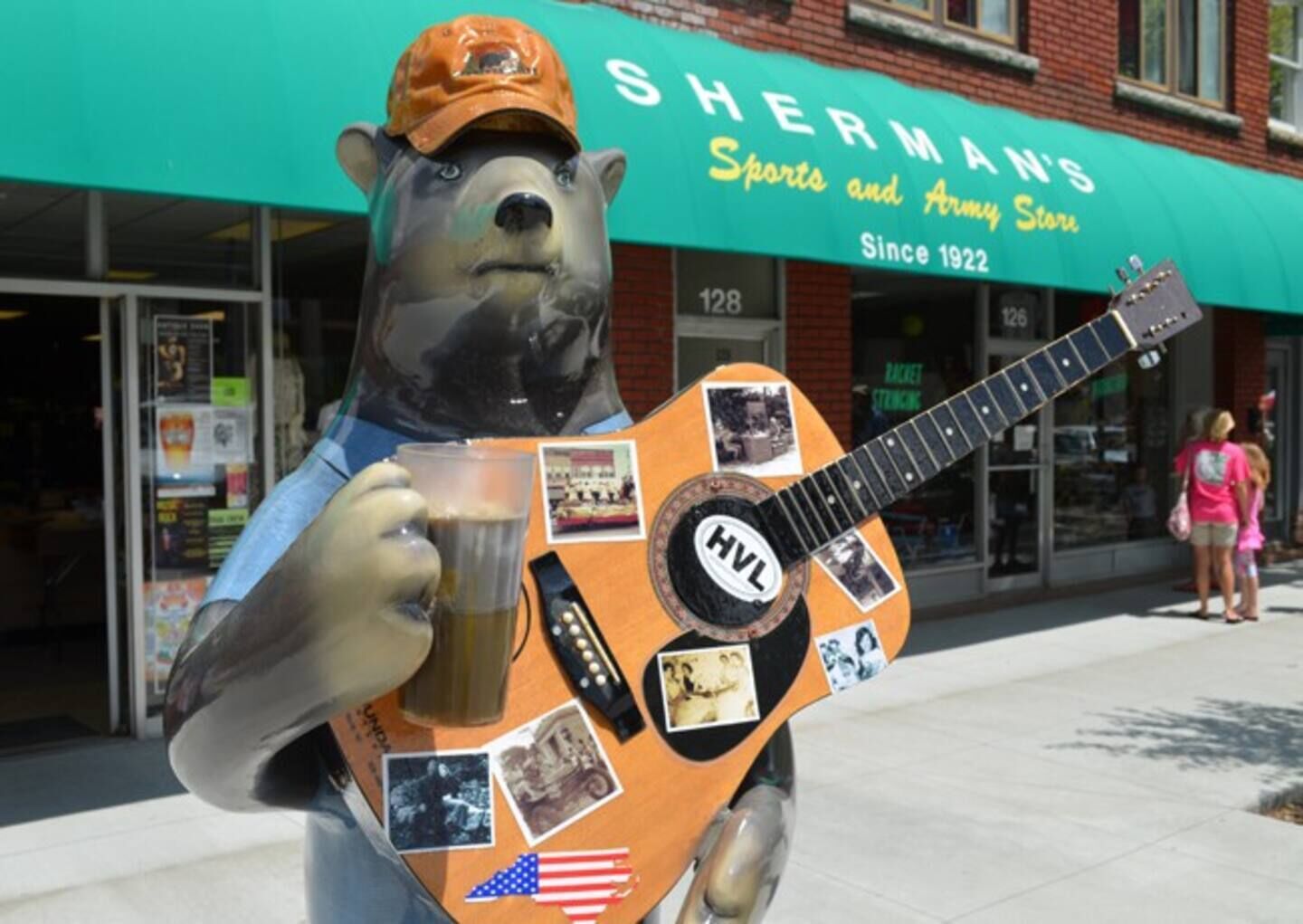 A bear statue holding a guitar and a mug in front of a sports and army store.