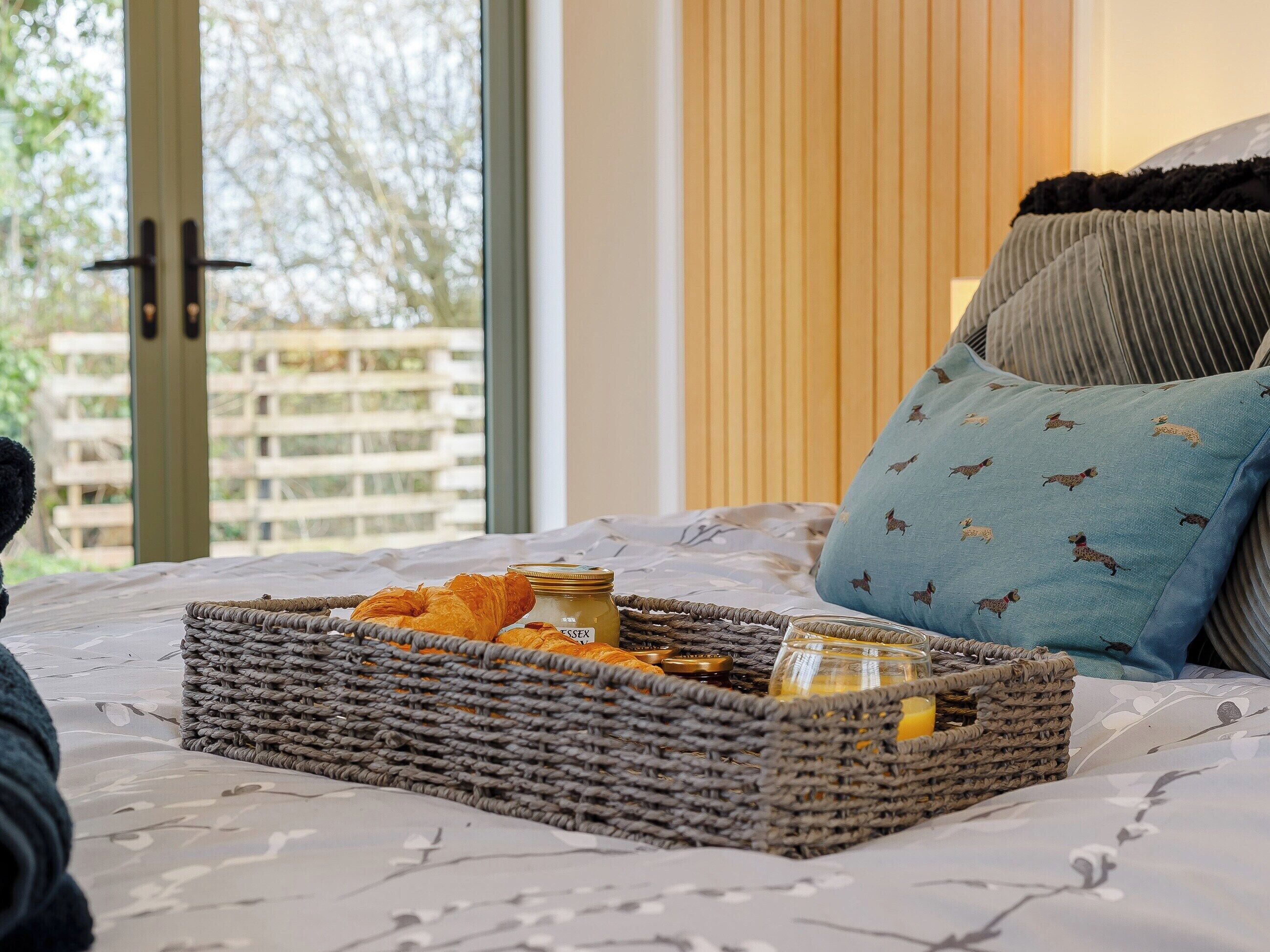 Un lit avec un panier en osier contenant des aliments, un oreiller orné d’un motif de chien et une vue sur une clôture en bois à l’extérieur.