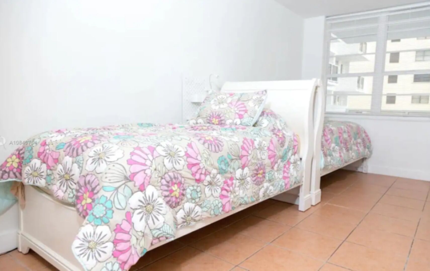 Un lit simple avec une couvre-lit à motifs floraux, dans une pièce aux murs blancs et dotée d’une fenêtre.
