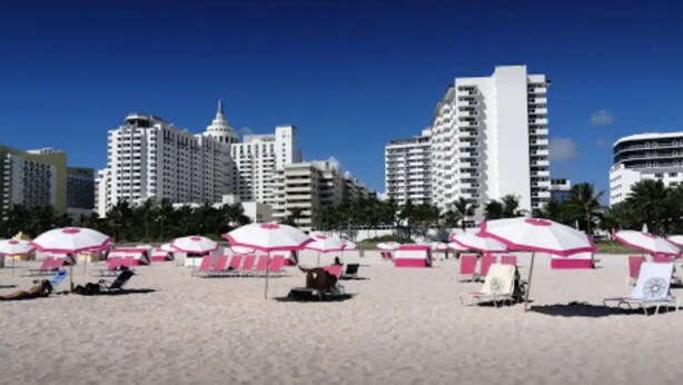 Una platja amb ombrelles roses i seients de platja, un cel blau clar i edificis alts al fons.