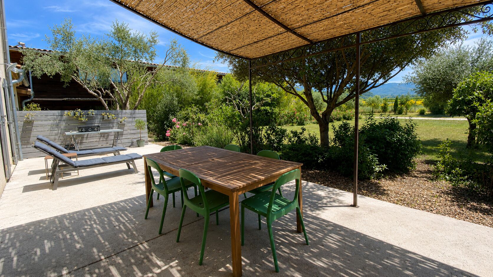 Un patio al aire libre con mesa de madera y sillas verdes, una parrilla y una pérgola que ofrece sombra.