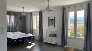 Ein Schlafzimmer mit einem Bett, zwei Pendelleuchten, einem Nachttisch, einem gerahmten Bild und großen Fenstern mit Vorhängen.
