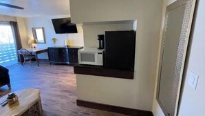 Hotellihuone, jossa on pieni keittiö, seinälle asennettu taulutelevisio, ruokailutila pöydällä ja tuoleilla sekä suuri ikkuna, josta tulee runsaasti luonnonvaloa.