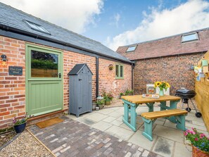 A brick house with a green door, a grey shed, and a patio with a table and bench.