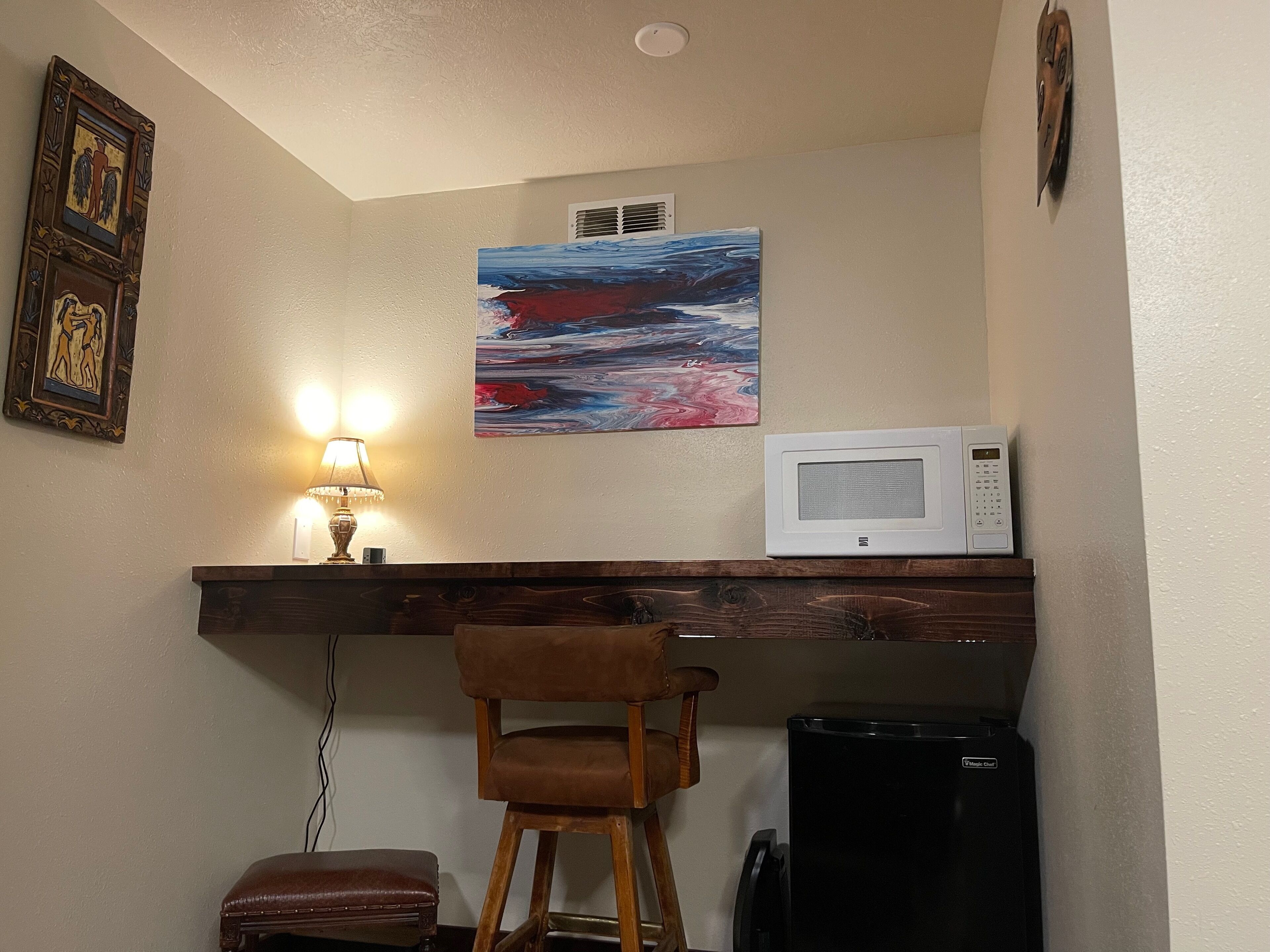 A compact kitchen with a microwave on a wooden shelf, a stool, and a trash bin.