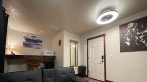 Een woonkamer met een zwarte bank, een witte magnetron, een kleine tafel met een lamp, een schilderij en een deur met een spiegel.