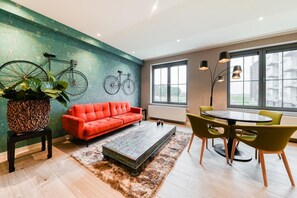 A modern living room with a red sofa, a coffee table, and a dining area with chairs. There are bicycles hanging on the wall and a large window providing natural light. - Luxury 1-bedroom apartment at coast's sportiest park (Middelkerke)