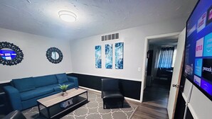 A living room with a blue sofa, a coffee table, and two black chairs. There are abstract paintings on the wall and a flat-screen TV displaying various channels. - Colorful Shadow-Game Rm, Hot Tub, 5 min from Chisholm Creek (Oklahoma City)