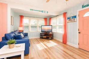 A living room with a blue sofa, a TV on a wooden stand, a ceiling fan, and a door labeled 'Beach'. - Saltwater Pool, 1 Min to Beach, Dog Friendly, Large Deck with Ocean Views 4BR3Ba (North Myrtle Beach)