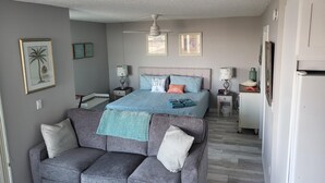Une chambre à coucher avec un lit, un canapé gris, un ventilateur de plafond et un réfrigérateur blanc.