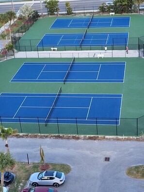 Et tennisanlæg med flere baner, omgivet af hegn, beliggende ved siden af en parkeringsplads med flere biler.