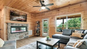Ein gemütliches Blockhausschlafzimmer mit Kamin aus Stein, Deckenventilator, grauem Sofa und großem Fenster mit Blick ins Freie.
